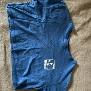 Blå t-shirt med tryck - Säljer en blå t-shirt med ett litet vitt tryck framtill. T-shirten har rund hals och korta ärmar, croppad modell, fin blå färg! Använd enstaka gång.