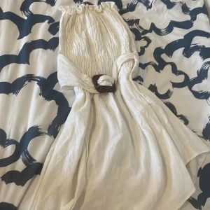 Vit klänning med bälte - Säljer en vit långklänning med smockad överdel och markerad midja med ett snyggt bälte i trä-look. Klänningen har en luftig och somrig känsla, perfekt för varma dagar.