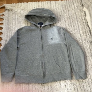 Grå hoodie från Polo Ralph Lauren - Klassisk grå hoodie från Polo Ralph Lauren med dragkedja och huva. Tröjan har två fickor framtill och en liten broderad logga på bröstet. Perfekt för en avslappnad och stilren look.