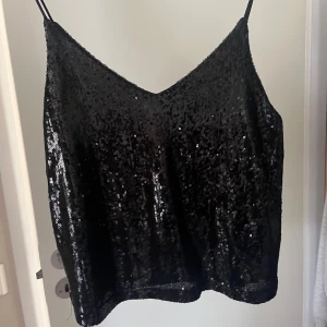 Svart paljettlinne med smala axelband - Säljer en svart topp med smala axelband och glittriga paljetter över hela toppen. Perfekt nu till våren och sommaren. Snygg v-ringning och lätt croppad passform. Den är helt oanvänd med prislappen kvar! Hör av er vid frågor 🥰