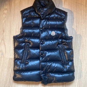 Moncler gilet Väst - Snygg svart dunväst från Moncler med hög krage och tryckknappar framtill.  köptes på nk i 2023 men har ej använts så mycket. Kvitto har tyvärr kastats bort. STORLEK S. Scan funkar för äktenhet.  kan mötas i Stockholm eller frakt. 