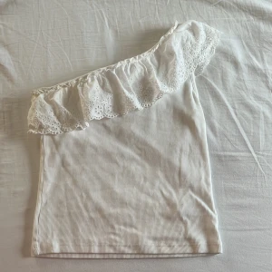 Vit one shoulder topp med volang - Säljer en vit ribbad one shoulder topp med bred volangkant i spets. Toppen är ärmlös och har en romantisk känsla med sin dekorativa volang. Perfekt för dig som vill sticka ut lite extra!