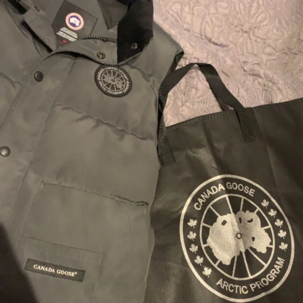Säljer en grå dunväst från Canada Goose med klassisk logotyp på bröstet och knappar framtill. Västen har två stora fickor och är perfekt för lager-på-lager. Stilsäker och varm för kyliga dagar byte kan tas imot hör av sig vid frågor eller intresse!. Takit.