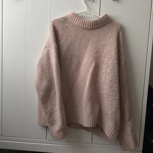 Beige/rosa stickad tröja från H&M - Mysig beige/ljus rosa stickad tröja från H&M med hög krage och långa ärmar. Tröjan har en avslappnad passform och breda ribbade muddar vid hals, ärmslut och nederkant. Perfekt för kyliga dagar eller en casual look.