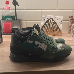 Valentino Rockrunner sneakers i grön camouflage - Säljer ett par Valentino Rockrunner sneakers i grönt med camouflage-mönster. Skorna har snörning, rund tå och är tillverkade i en mix av mocka, skinn och textil. Perfekta för dig som vill sticka ut med en exklusiv streetwear-look. Skriv innan köp färg har gått bort lite men går att måla tillbaka 🤩