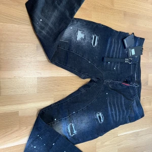 Dsquared2 Jeans - Säljer ett par svarta jeans från Dsquared2 med slitna detaljer och färgstänk. Jeansen har flera revor och en klassisk femficksmodell. Märkets logga syns på en röd etikett vid fickan och medföljande tagg.