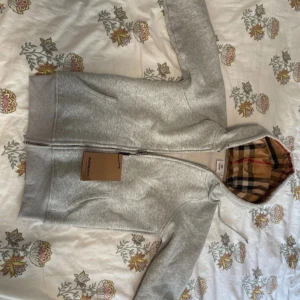 HELT NY BURBERRY HOODIE - Säljer en grå hoodie från Burberry i storlek M men passar som S. Tröjan har dragkedja framtill, fickor och den klassiska Burberry-rutiga fodret i huvan. Perfekt för dig som gillar stilrena och exklusiva plagg.