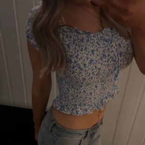 Blommig croppad blus med volangkant - Söt blommig croppad blus i vitt och blått med volangkant och rosett fram till. Offshoulder-modell med korta ärmar och smockad resår upptill och nertill. Perfekt för varma dagar!