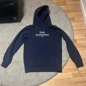 Mörkblå hoodie från Peak Performance - Säljer en mörkblå hoodie från Peak Performance med vit logga på bröstet. Tröjan har huva och en stor magficka framtill. Perfekt för dig som gillar en klassisk och stilren look.