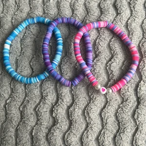 Färgglada armband med hjärtdetalj - Tre elastiska armband i blått, lila och rosa med platta pärlor. Ett armband har en vit pärla med rosa hjärta. Perfekta för att addera färg till din stil!