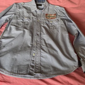 Randig skjorta från Napapijri - Säljer en blåvit randig skjorta från Napapijri med två bröstfickor och broderad norsk flagga på vänster sida. Skjortan har klassisk krage, knappar framtill och axelklaffar. Perfekt för dig som gillar stilrena detaljer.