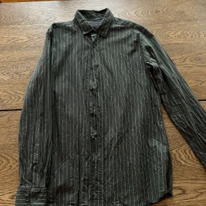 Grön randig skjorta från Massimo Dutti - Snygg grön skjorta med vita ränder från Massimo Dutti. Skjortan är långärmad och har klassisk krage samt knäppning framtill. Tillverkad i 100% bomull för en skön känsla och stilren look. Storlek L, använd en gång. 