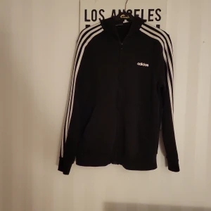 Svart Adidas track jacket med vita ränder - Klassisk svart Adidas track jacket med vita ränder längs ärmarna och dragkedja framtill. Jackan har hög krage och Adidas-logga på bröstet. Perfekt för en sportig och avslappnad stil.