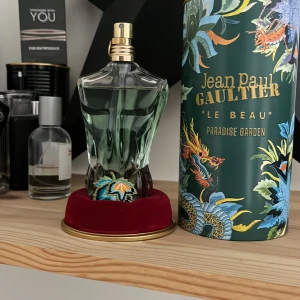 Jean Paul Gaultier Le Beau Paradise Garden Eau de Parfum - Parfymflaska formad som en manlig torso i grönt glas med guldfärgad kork. Kommer med en dekorativ grön förpackning med tropiskt mönster och texten 'Le Beau Paradise Garden'.
