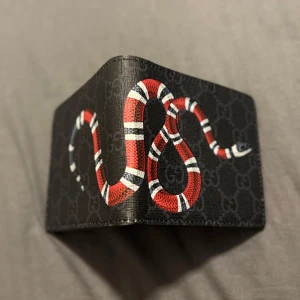 Svart plånbok med orm från Gucci - Snygg svart plånbok från Gucci med ikoniskt ormmotiv i rött, vitt och svart på utsidan. Plånboken har flera kortfack, fönster för ID och diskret GG-mönster i bakgrunden. Perfekt accessoar för dig som gillar lyxiga detaljer.
