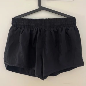 Svarta träningsshorts med resår och snörning - Svarta träningsshorts med bred resår i midjan och snörning på insidan av shortsen för extra passform. Lätta och luftiga med innerbyxa, perfekt för sport eller gym. Klassisk design utan mönster och med diskret logga bak. Köpta i USA och aldrig använda