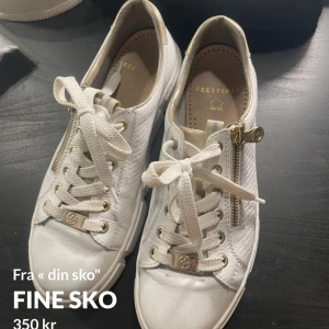 Vita sneakers från Feetfirst med dragkedja - Säljer ett par vita sneakers från Feetfirst med snörning och guldfärgade detaljer. Skorna har dragkedja på sidan och en diskret mönstrad textur på sidorna. Perfekta för en stilren och fräsch look.