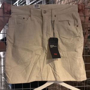Vit manchesterkjol från Levi's - Säljer en snygg vit manchesterkjol från Levi's med klassisk femficksdesign och knappgylf. Kjolen har en rak passform och är tillverkad i mjuk manchester, perfekt för en trendig vardagslook. Aldrig använd med lappar kvar! 