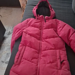 Röd pufferjacka med huva från Take Off - Säljer en röd pufferjacka från Take Off med huva och dragkedja framtill. Jackan har två fickor och är vadderad för extra värme. Perfekt för kyliga dagar och enkel att matcha med olika outfits.