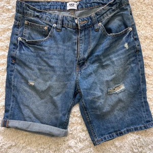 Blå jeansshorts Stl S - Säljer ett par klassiska blå jeansshorts från Laget 157 med slitna detaljer och fem fickor. Modellen har normal passform och uppvikta benslut. Mycket bra skick, använda en eller två gånger.