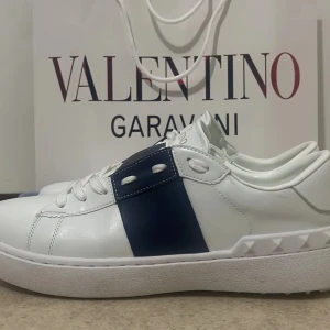 Valentino Garavani vita sneakers med blå detalj - Snygga vita sneakers från Valentino Garavani med en bred blå remsa på sidan och ikoniska nitar på hälen. Skorna har rund tå, platt sula och är tillverkade i skinn. Perfekta för dig som vill ha en stilren och lyxig look.