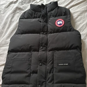 Svart dunväst från Canada Goose - Hej Säljer en äkta svart dunväst från Canada Goose med klassisk logotyp på bröstet och två stora fickor framtill. Västen har hög krage och stängs med knappar. Perfekt för lager-på-lager under kyliga dagar. Den e i storlek xs men passar s skriv vid fler funderingar (äkta väst) pris ej hugget i sten 