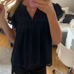 Mörkblå blus med volangdetaljer - Snygg mörkblå blus med volangdetaljer på ärmarna och v-ringad hals. Perfekt för en stilren look med en touch av elegans.
