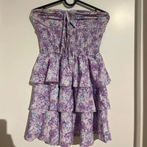 Lila blommig volangklänning - Supersöt kort klänning med lila och vita blommor, volanger i flera lager och smock upptill. Kan bäras axelbandslös eller med smala axelband tack vare knytningen. Perfekt för sommarens alla tillfällen!