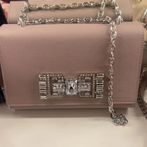 Rosa axelväska med kedja och strassdetalj - Säljer en stilren rosa axelväska med silverfärgad kedja och dekorativt spänne med stora strasstenar framtill. Väskan har en elegant och boxig form, perfekt för att lyfta din outfit. Använd en gång, inga defekter. Köptes för ca 500kr