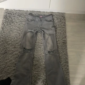 Grå slitna jeans - Säljer ett par gråa jeans med slitna detaljer och hål på båda benen. Byxorna har en bootcut passform och klassisk femficksdesign. De passar mig som har 34 i byxor. Använda fåtal gånger.