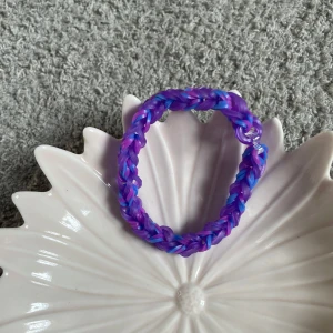 Färgglatt armband i lila och blått - Säljer ett handgjort armband i elastiska gummiband med flätad design i lila, blå och rosa nyanser. Perfekt accessoar för att addera lite färg till din stil!