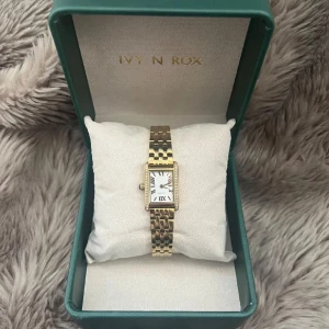 Klocka - Elegant guldfärgad smyckesklocka från Ivy N Rox med kvartsurverk. Klockan har ett rektangulärt ur med romerska siffror och ett metallband i rostfritt stål. Perfekt accessoar för att lyfta din stil.