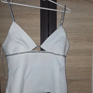 Vit topp med glittriga detaljer - Snygg vit linnetopp med smala, glittriga axelband och en dragkedja i ryggen. Perfekt för en kväll ute eller en festlig tillställning. Aldrig använd och prislappen är på. 