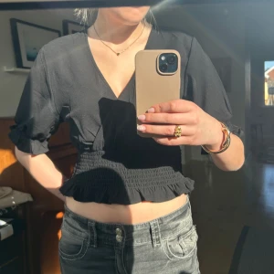 Svart blus från Ginatricot - Snygg svart omlottopp från Ginatricot med v-ringning och lite kortare ärmar. Toppen har smockdetaljer i midjan och volangkant. Endast använd ett par fåtal gångar. 