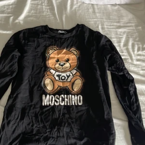 Svart långärmad tröja från Moschino - Säljer en svart långärmad tröja från Moschino med en stor nallebjörn och texten 'Toy' på framsidan. Tröjan har en rund halsringning och är perfekt för en avslappnad stil. Passar bra till jeans eller kjol för en cool look.