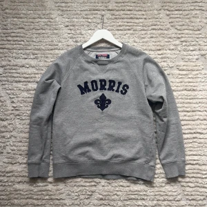 Grå sweatshirt från Morris - Säljer en stilren grå sweatshirt från Morris med mörkblå text och emblem på framsidan. Tröjan har långa ärmar och rund halsringning. Skick: 7/10. Storlek: (M)