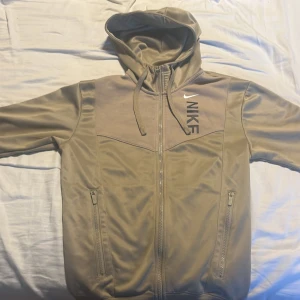 Beige huvtröja från Nike - Säljer en beige huvtröja från Nike med dragkedja och justerbar huva. Tröjan har långa ärmar och två fickor med dragkedjor framtill. Perfekt för en avslappnad stil eller träning. Storleken är xs men kan passa S också 