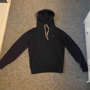 Marinblå  hoodie från Ralph Lauren - Säljer en stilren svart hoodie från Ralph Lauren med klassisk design och dragsko i huvan. Perfekt för en avslappnad look. Den har långa ärmar och en liten röd logga på bröstet. Passar perfekt för kyliga dagar.