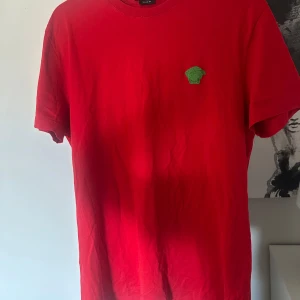 Röd t-shirt från Versace - Säljer en snygg röd t-shirt från Versace med ett grönt broderat märke på bröstet. T-shirten är kortärmad och har en klassisk passform. Perfekt för dig som vill ha en stilren look med en touch av lyx.