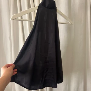 Svart halterneck-topp med knytband - Elegant svart halterneck-topp med knytband i nacken. Perfekt för en stilren look. Tillverkad i ett glansigt material som ger en lyxig känsla. Aldrig kommit till användning då jag inte tycker den ser bra ut på mig. 