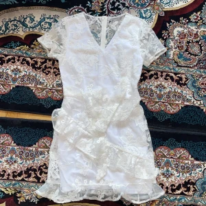 Vit spetsklänning med volangdetaljer - Säljer en elegant vit klänning med v-ringning och korta ärmar. Klänningen har vackra spetsdetaljer och volanger som ger en romantisk känsla. Perfekt för speciella tillfällen så som studenten🥰