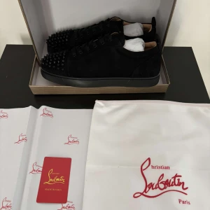 Svarta sneakers med nitar från Christian Louboutin - Snygga svarta sneakers i mocka från Christian Louboutin med ikoniska röda sulor. Skorna har nitar på tån och snörning för en edgy look. Perfekta för att ge din outfit en lyxig touch.