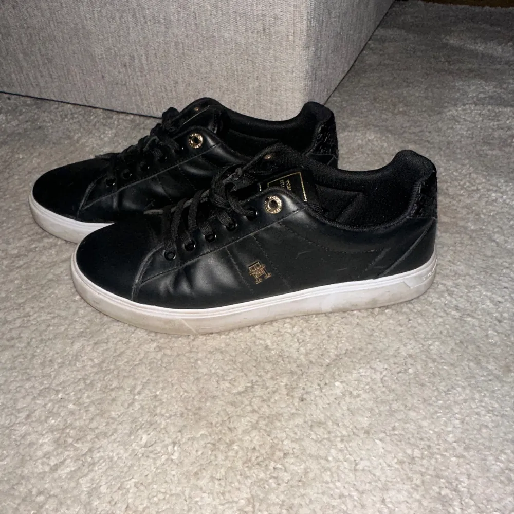 Snygga svarta sneakers med gulddetaljer och snörning. Skorna har en vit sula och en diskret logga på sidan. Perfekta för en stilren look.. Kengät.
