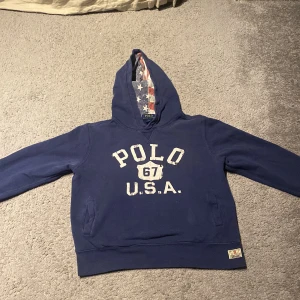 Blå hoodie från Ralph Lauren - Säljer en blå hoodie från Ralph Lauren med texten 'POLO 67 U.S.A.' på framsidan. Huvan är fodrad med ett mönster av den amerikanska flaggan. Perfekt för en avslappnad stil.