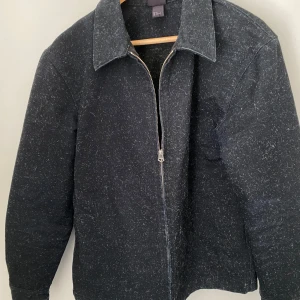 Svart jacka med dragkedja från H&M - Snygg svart jacka med dragkedja från H&M och klassisk krage. Jackan har långa ärmar och knappar vid ärmsluten för en stilren look. Perfekt för både höst och vår.