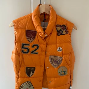 Orange dunväst från Moncler - Säljer en cool orange dunväst från Moncler med flera unika patches och ett stort 'M' på ryggen. Västen har knappar framtill och en hög krage för extra stil och värme. Perfekt för att sticka ut i mängden!