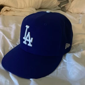 Blå keps med LA-logga - Snygg blå keps från New Era med LA-logga framtill. Kepsen har en klassisk design med böjd skärm och broderad logga på sidan. Perfekt för att komplettera din streetwear-look. Kepsen är i storlek 58,7