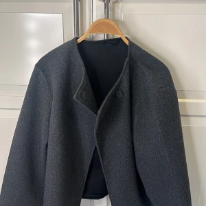 Grå kappa från Gina Tricot - Snygg grå kappa från Gina Tricot med en enkel knapp framtill. Den har en stilren design och är perfekt för kyligare dagar. Passar bra till både vardag och mer uppklädda tillfällen.