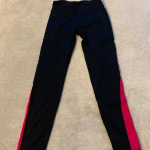 Svarta träningsleggings med rosa detaljer - Snygga svarta träningsleggings med rosa sidopaneler och reflexdetaljer. Perfekta för löpning eller gymmet. De har en tight passform och är tillverkade i ett stretchigt syntetmaterial för bästa komfort.