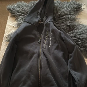 Svart Hoodie från Sail Racing - Snygg svart hoodie från Sail Racing med dragkedja och huva. Hoodien har en praktisk bröstficka med dragkedja och justerbar huva för extra komfort. Perfekt för kyligare dagar. Och Dragkedjan är av på bröstfickan. 